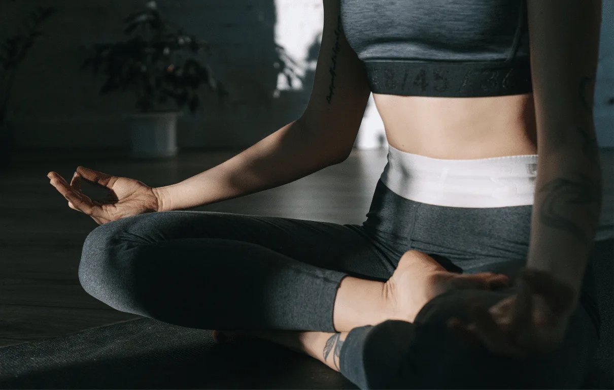 Woman in gray activewear seated cross-legged in meditation on a studio floor.