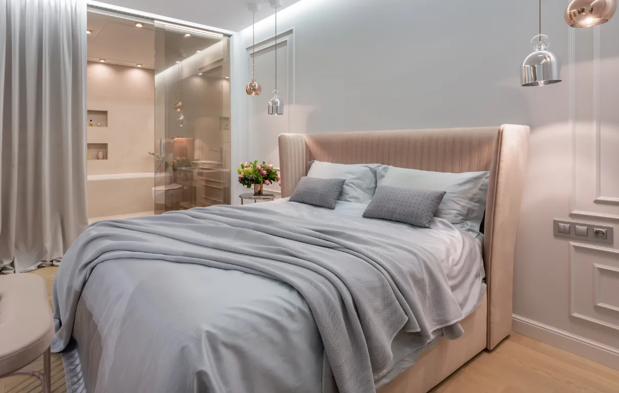 A softly lit bedroom with layered pastel bedding and a blush upholstered headboard.